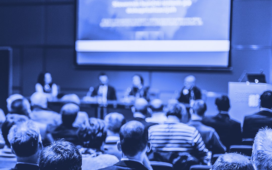 A conference audience facing a panel on stage beneath a large presentation screen, tinted blue